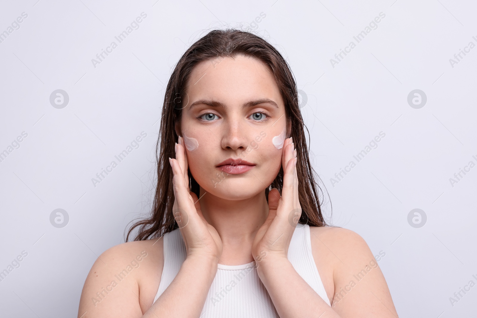 Photo of Beautiful woman applying skincare product onto her face on light grey background, closeup