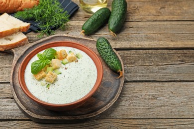 Photo of Tasty cucumber soup with croutons, basil, bread and vegetables on wooden table. Space for text
