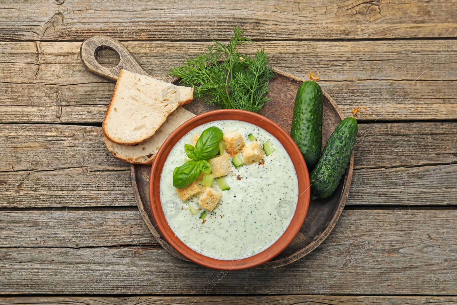 Photo of Tasty cucumber soup with croutons, basil, bread and vegetables on wooden table, flat lay