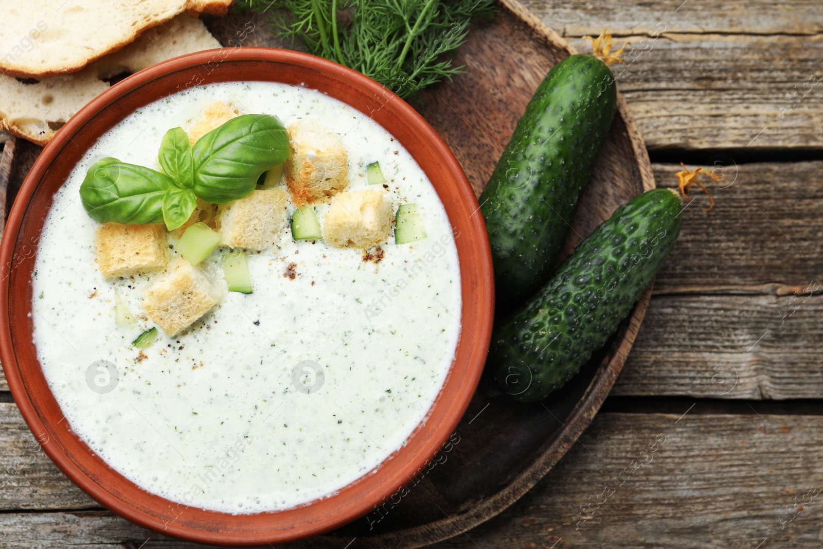 Photo of Tasty cucumber soup with croutons, basil, bread and vegetables on wooden table, flat lay