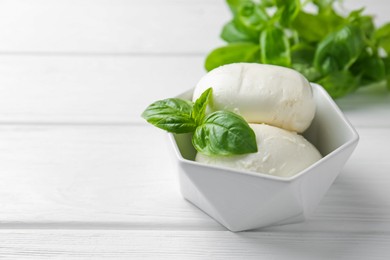 Photo of Pieces of delicious mozzarella cheese and basil in bowl on white wooden table, closeup. Space for text