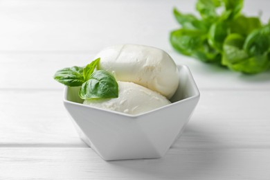 Photo of Pieces of delicious mozzarella cheese and basil in bowl on white wooden table, closeup