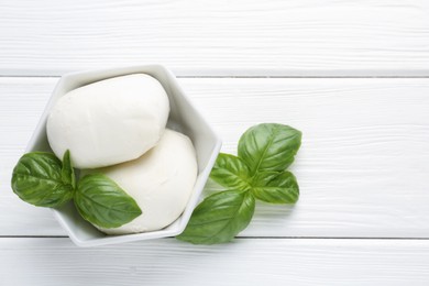 Photo of Pieces of delicious mozzarella cheese and basil in bowl on white wooden table, flat lay. Space for text