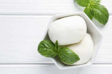 Photo of Pieces of delicious mozzarella cheese and basil in bowl on white wooden table, flat lay. Space for text