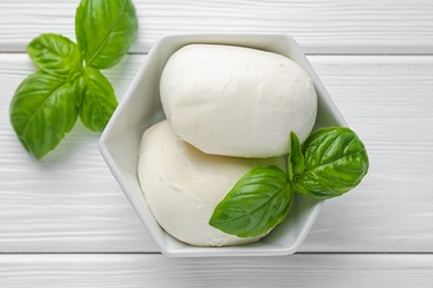 Photo of Pieces of delicious mozzarella cheese and basil in bowl on white wooden table, flat lay