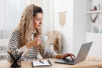 Photo of Woman with cup of hot drink working on laptop at desk in home office