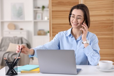 Photo of Smiling woman talking by smartphone with laptop at desk in home office