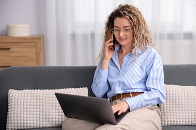 Photo of Smiling woman with laptop talking by smartphone on sofa in home office