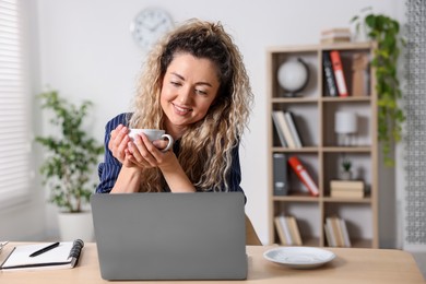 Photo of Smiling woman with cup of hot drink working on laptop at desk in home office