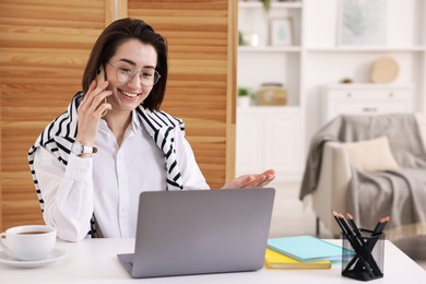 Photo of Smiling woman talking by smartphone with laptop at desk in home office