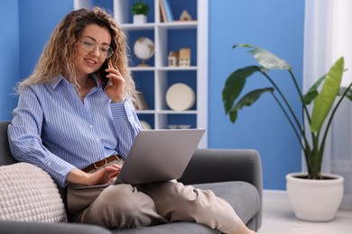 Photo of Smiling woman with laptop talking by smartphone on sofa in home office