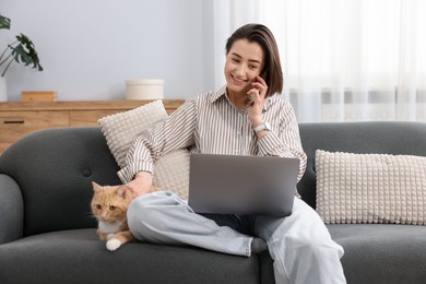 Photo of Smiling woman talking by smartphone with laptop and cat on sofa in home office