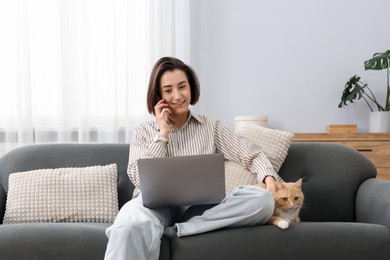 Photo of Smiling woman talking by smartphone with laptop and cat on sofa in home office