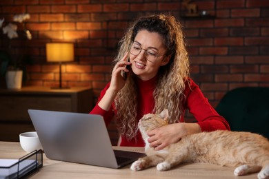 Photo of Smiling woman talking by smartphone and stroking cat at desk in home office