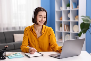 Photo of Smiling woman in headphones having videochat by laptop at desk in home office
