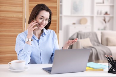 Photo of Smiling woman talking by smartphone with laptop at desk in home office