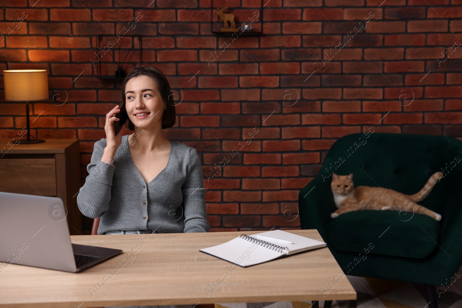 Smiling woman talking by smartphone at desk with laptop in home office Photo of Smiling woman talking by smartphone at desk with laptop in home office