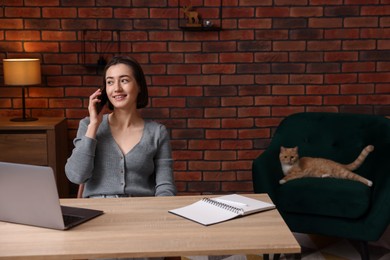 Photo of Smiling woman talking by smartphone at desk with laptop in home office