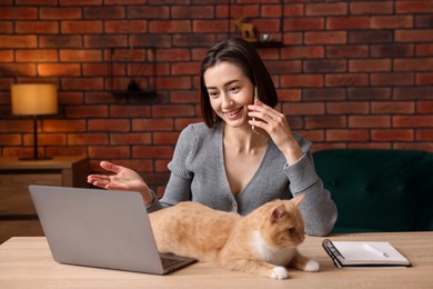 Photo of Smiling woman with cat talking by smartphone at desk in home office