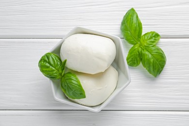 Photo of Pieces of delicious mozzarella cheese and basil in bowl on white wooden table, flat lay