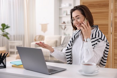 Photo of Smiling woman talking by smartphone with laptop at desk in home office