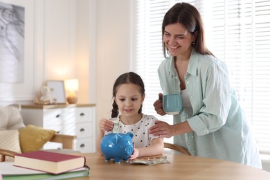 Photo of Mother and daughter putting pocket money into piggybank at table indoors