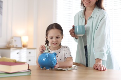 Photo of Mother and daughter putting pocket money into piggybank at table indoors, closeup