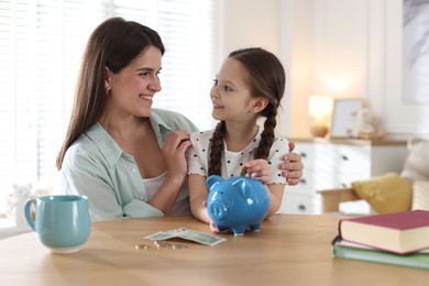 Photo of Mother and daughter putting pocket money into piggybank at table indoors