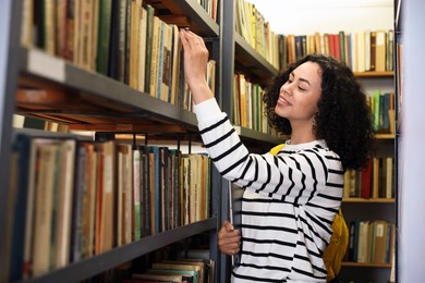 Photo of Woman with laptop choosing book from shelf in public library