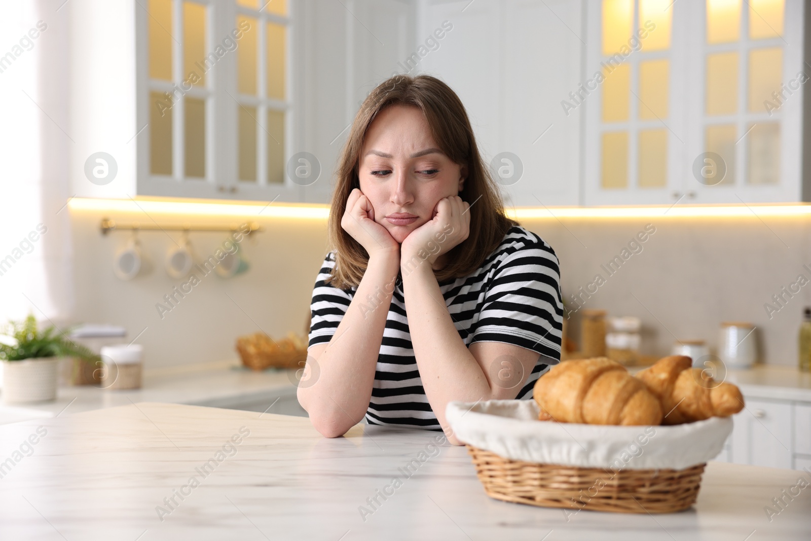 Gluten free diet. Sad woman looking at croissants at white marble table in kitchen Photo of Gluten free diet. Sad woman looking at croissants at white marble table in kitchen