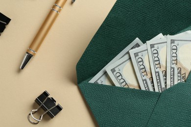 Photo of Money bonus. Envelope with dollar banknotes, pen and binder clip on beige background, flat lay