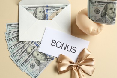 Photo of Card with word Bonus, envelope, dollar banknotes, bow and piggy bank on beige background, flat lay