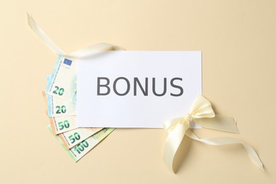 Photo of Card with word Bonus, euro banknotes and bow on beige background, flat lay