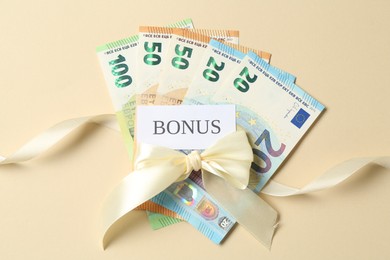 Photo of Card with word Bonus, euro banknotes and bow on beige background, flat lay
