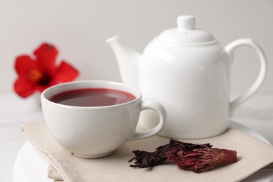 Photo of Delicious hibiscus tea in cup, teapot and dry roselle sepals on white table, closeup