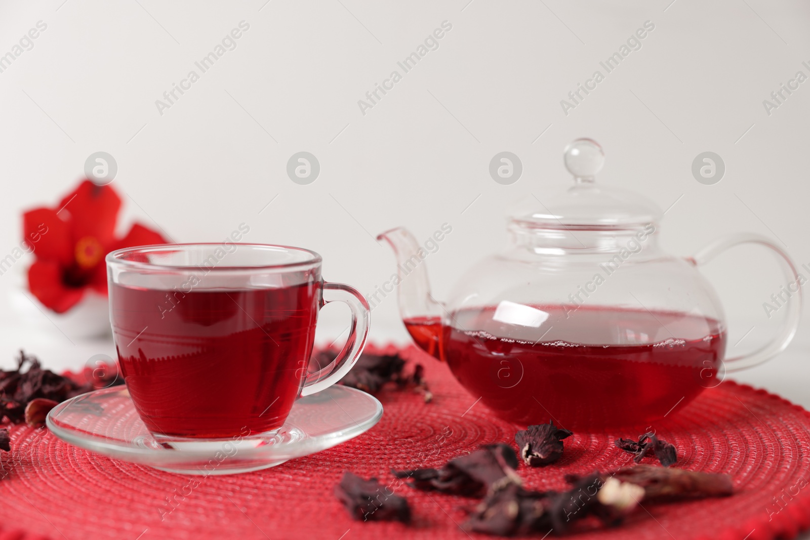Photo of Delicious hibiscus tea in glass cup, teapot and dry roselle sepals on red mat, closeup