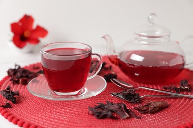 Photo of Delicious hibiscus tea in glass cup, teapot, dry roselle sepals and spoons on white table, closeup