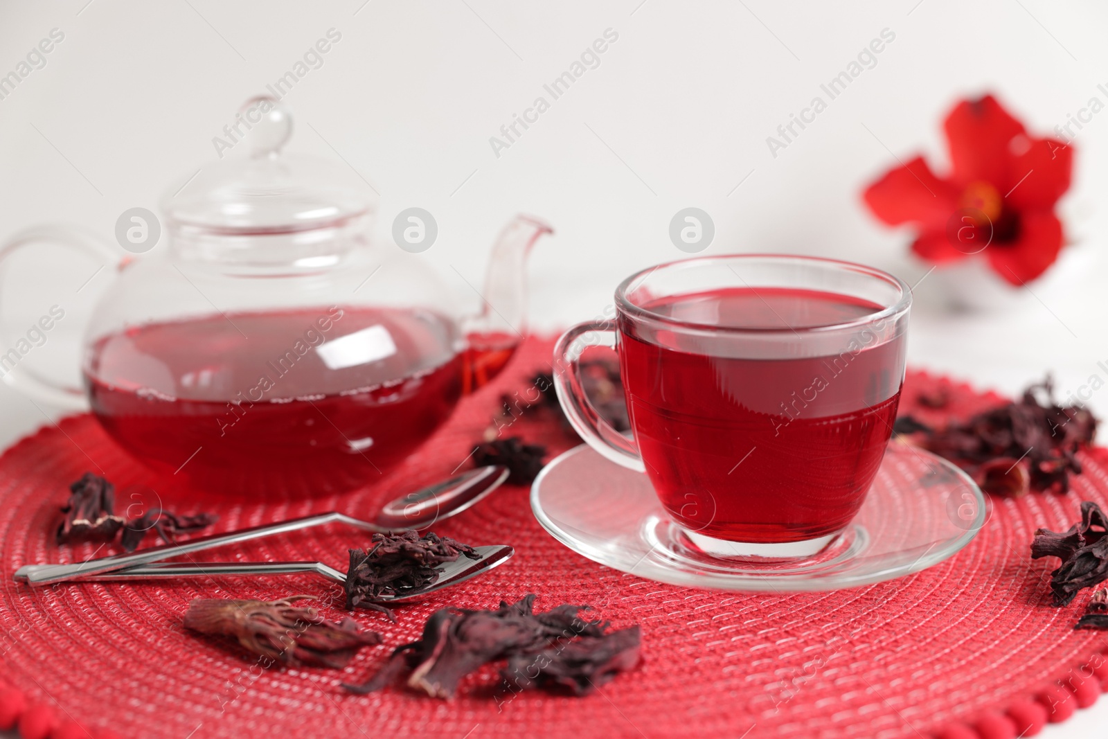 Delicious hibiscus tea in glass cup, teapot, dry roselle sepals and spoons on white table, closeup Photo of Delicious hibiscus tea in glass cup, teapot, dry roselle sepals and spoons on white table, closeup