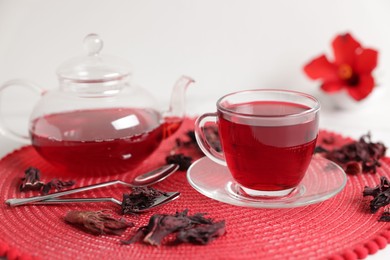 Photo of Delicious hibiscus tea in glass cup, teapot, dry roselle sepals and spoons on white table, closeup