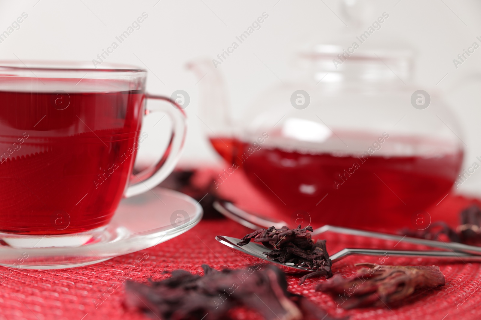 Delicious hibiscus tea in glass cup, teapot, dry roselle sepals and spoons on red mat, closeup Photo of Delicious hibiscus tea in glass cup, teapot, dry roselle sepals and spoons on red mat, closeup