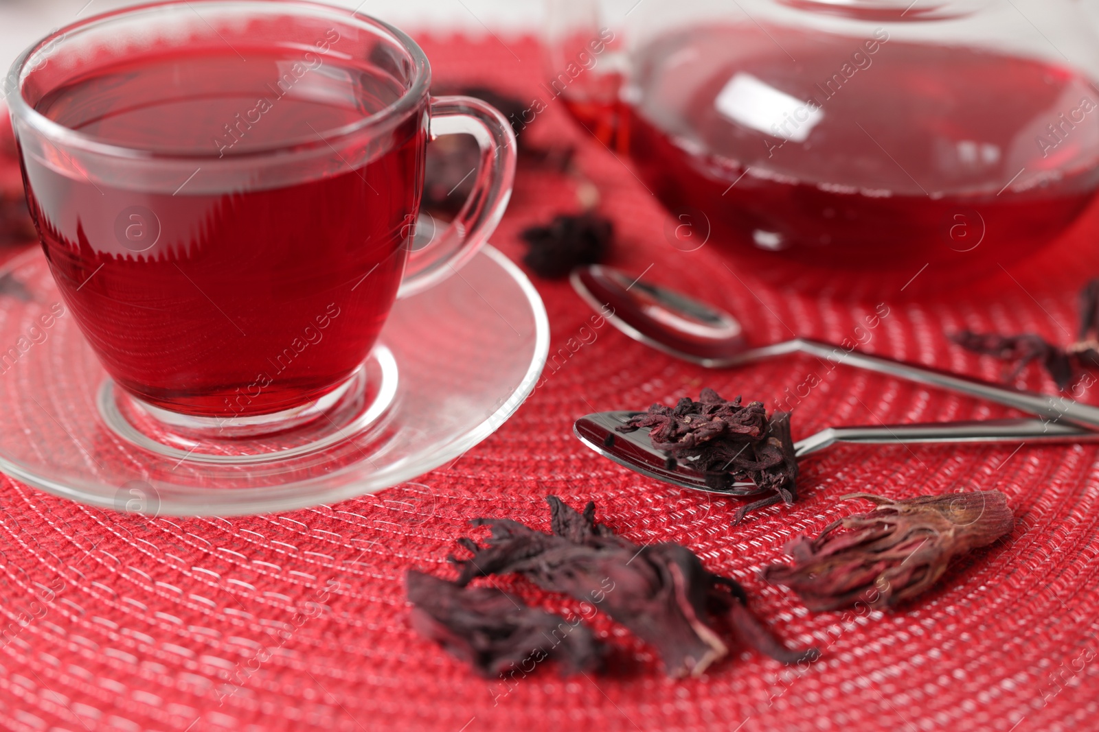 Photo of Delicious hibiscus tea in glass cup, teapot, dry roselle sepals and spoons on red mat, closeup