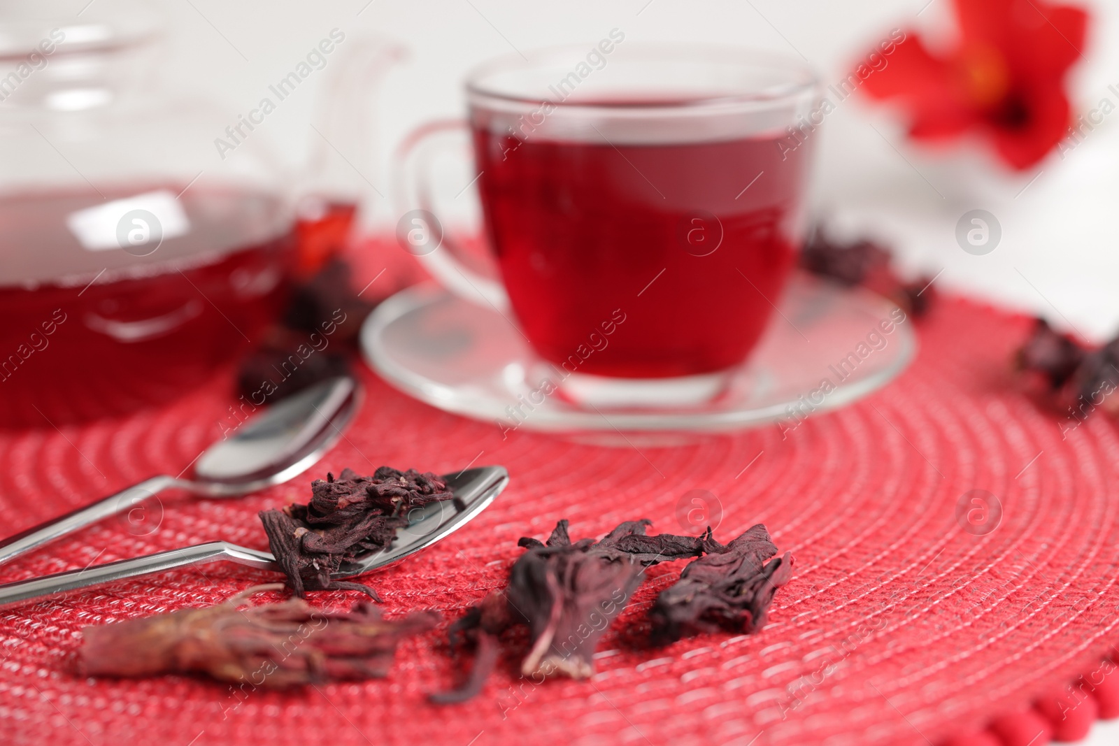 Delicious hibiscus tea in glass cup, teapot, dry roselle sepals and spoons on red mat, closeup Photo of Delicious hibiscus tea in glass cup, teapot, dry roselle sepals and spoons on red mat, closeup