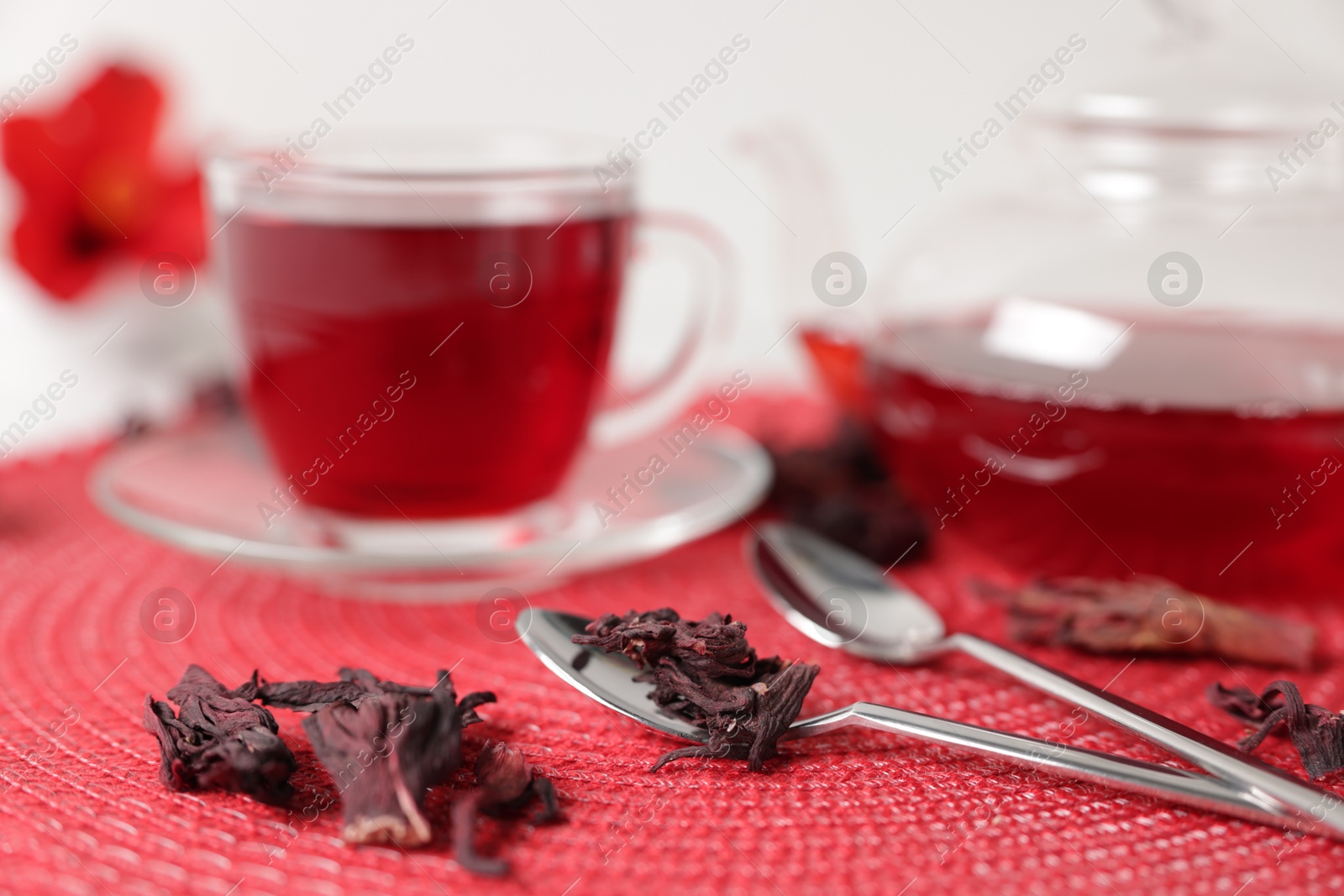 Delicious hibiscus tea in glass cup, teapot, dry roselle sepals and spoons on red mat, closeup Photo of Delicious hibiscus tea in glass cup, teapot, dry roselle sepals and spoons on red mat, closeup