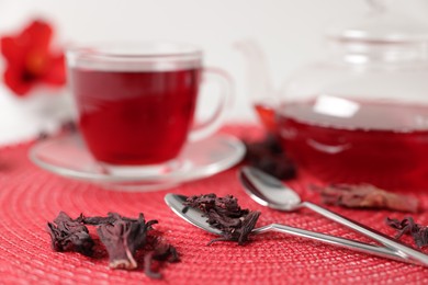 Photo of Delicious hibiscus tea in glass cup, teapot, dry roselle sepals and spoons on red mat, closeup