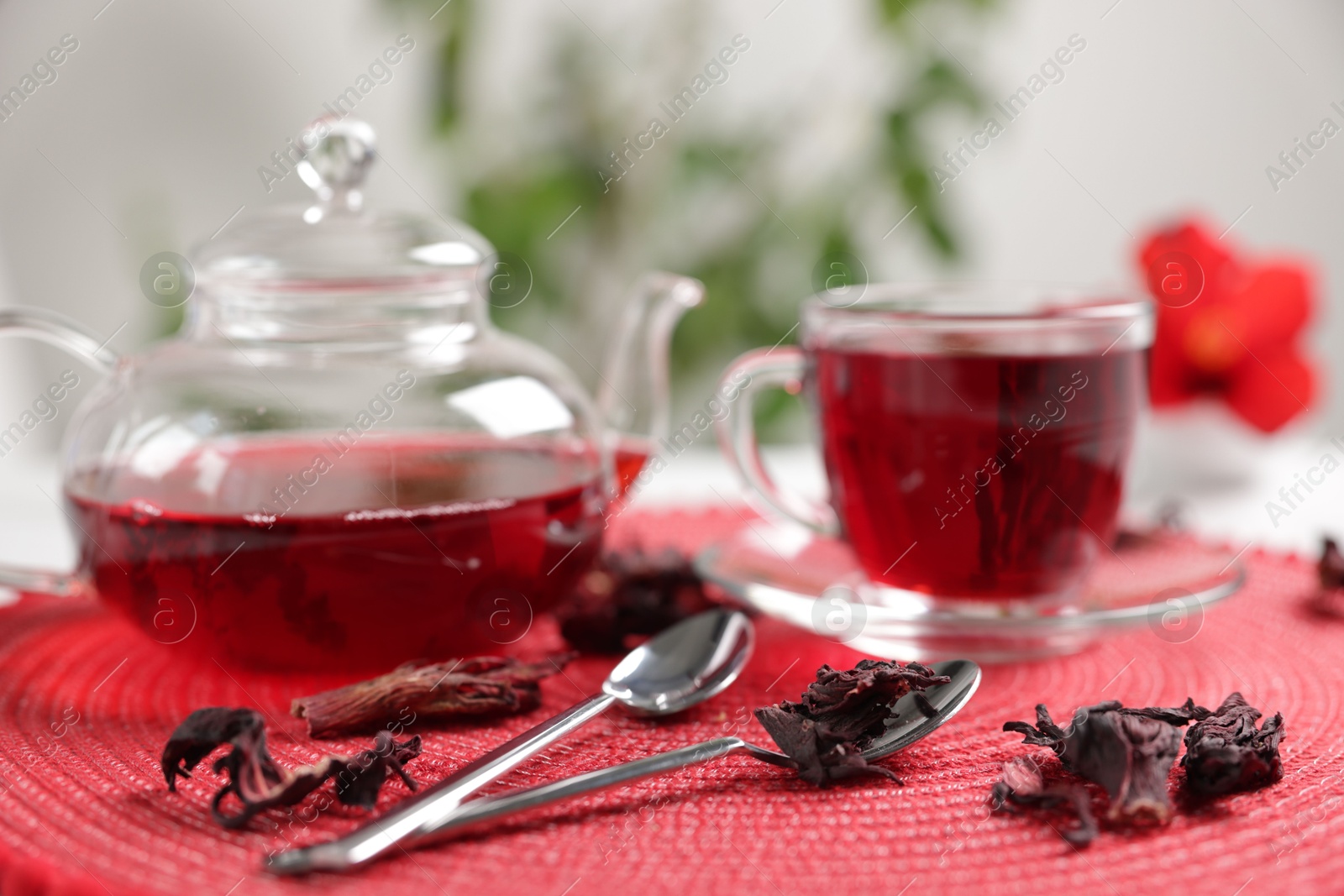 Delicious hibiscus tea in glass cup, teapot, dry roselle sepals and spoons on red mat, closeup Photo of Delicious hibiscus tea in glass cup, teapot, dry roselle sepals and spoons on red mat, closeup