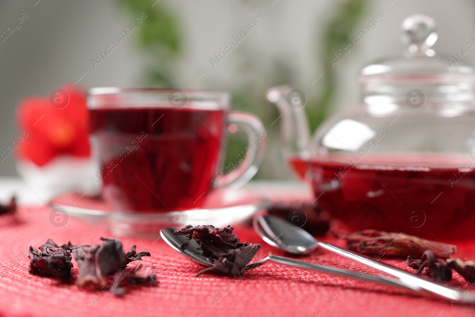 Delicious hibiscus tea in glass cup, teapot, dry roselle sepals and spoons on red mat, closeup Photo of Delicious hibiscus tea in glass cup, teapot, dry roselle sepals and spoons on red mat, closeup
