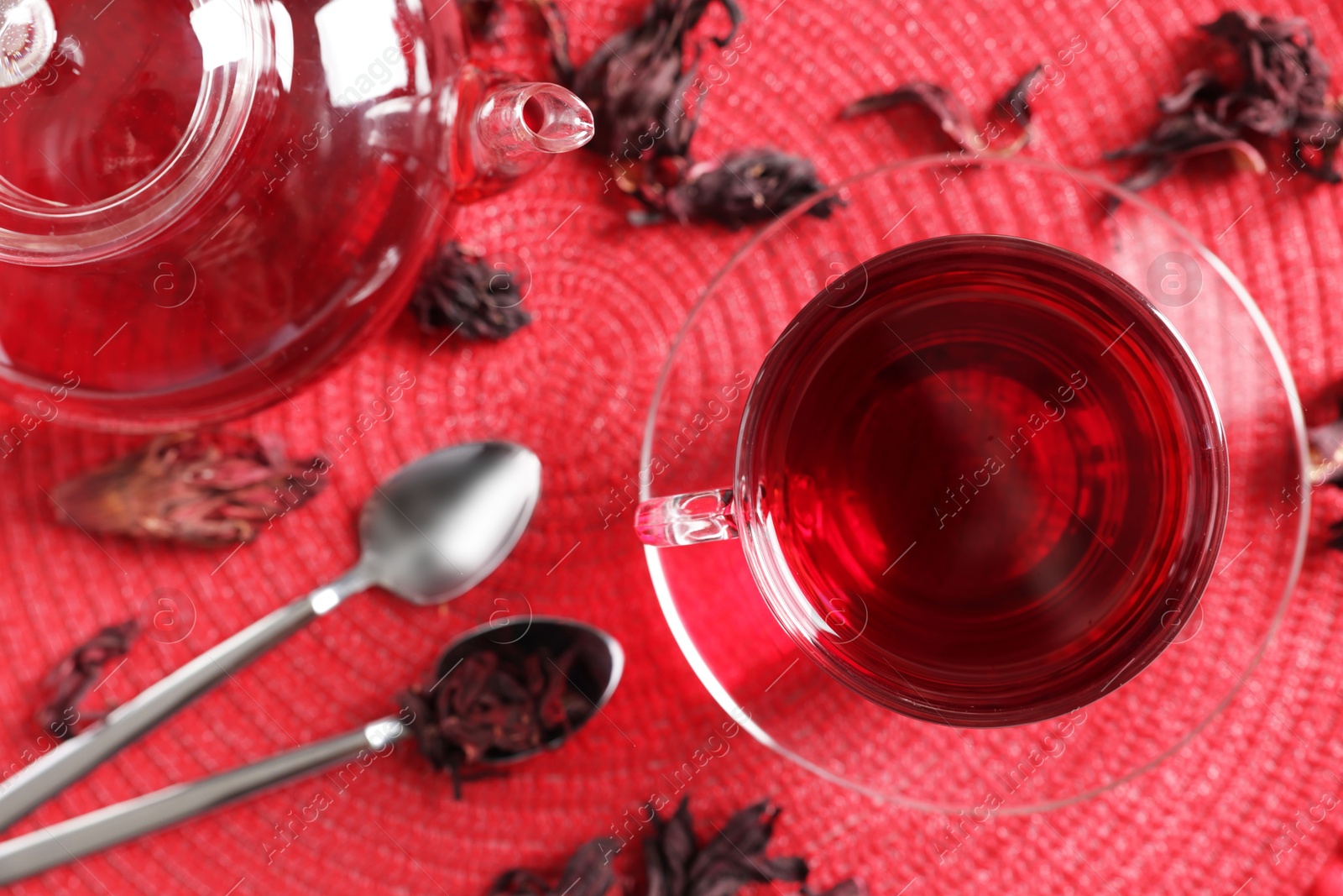 Delicious hibiscus tea in glass cup, teapot, dry roselle sepals and spoons on red mat, flat lay Photo of Delicious hibiscus tea in glass cup, teapot, dry roselle sepals and spoons on red mat, flat lay