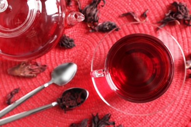 Photo of Delicious hibiscus tea in glass cup, teapot, dry roselle sepals and spoons on red mat, flat lay