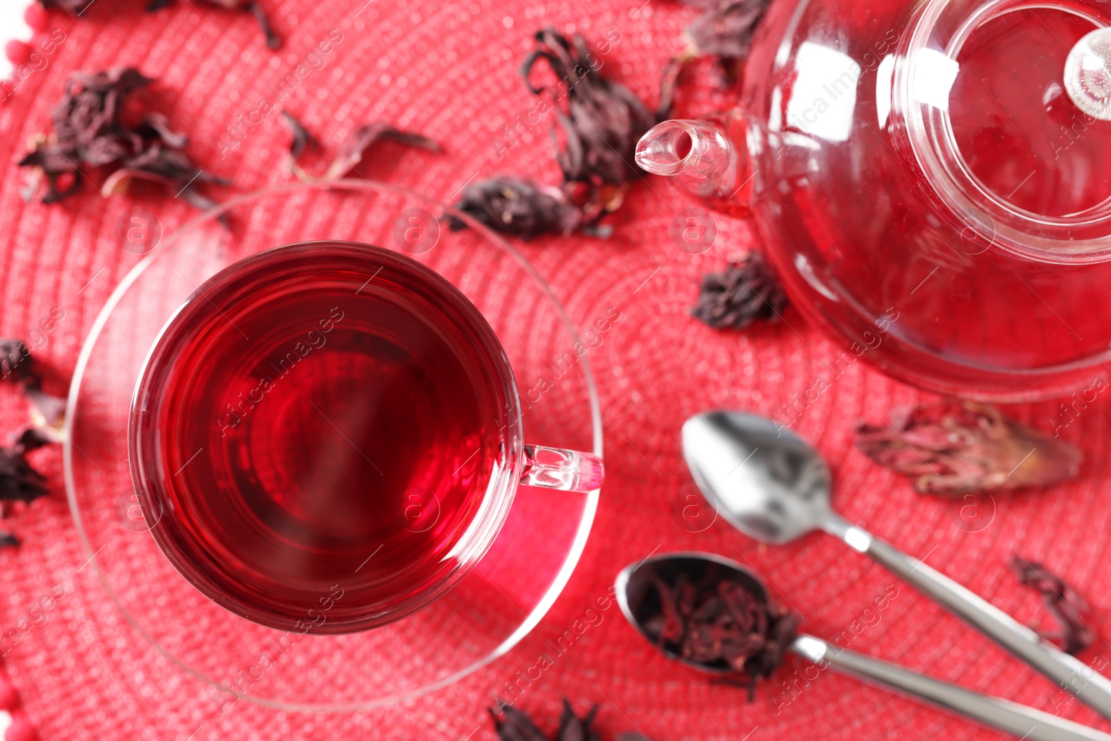 Delicious hibiscus tea in glass cup, teapot, dry roselle sepals and spoons on red mat, flat lay Photo of Delicious hibiscus tea in glass cup, teapot, dry roselle sepals and spoons on red mat, flat lay