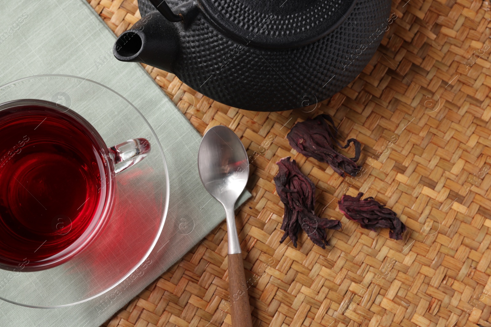 Delicious hibiscus tea in glass cup, teapot, dry roselle sepals and spoon on wicker mat, flat lay Photo of Delicious hibiscus tea in glass cup, teapot, dry roselle sepals and spoon on wicker mat, flat lay
